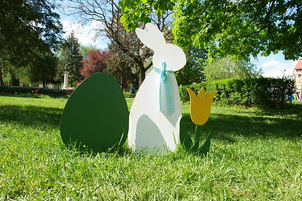 Easter bunny and egg decorations displayed on green grass in a sunny city park. Minimalist wooden rabbit figure with ribbon stands beside a large decorative egg and spring flower elements, surrounded by fresh foliage and trees. Outdoor seasonal setup celebrating Easter tradition, renewal, and joyful springtime atmosphere. Ideal for holiday themes, family celebrations, childhood concepts, and cheerful urban nature scenes.