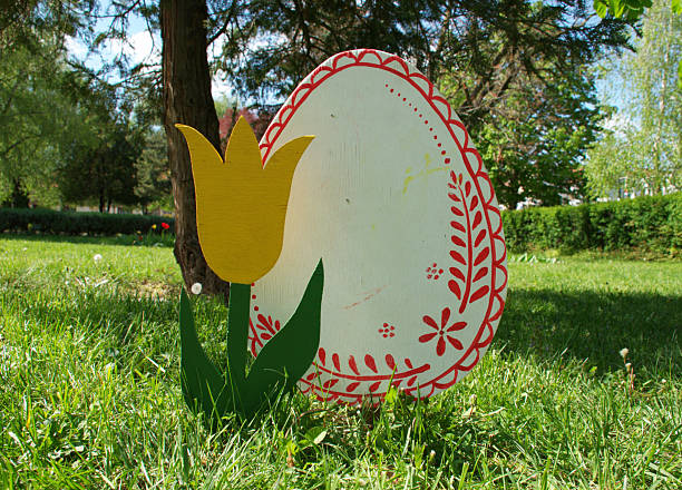 Decorative painted Easter egg with folk style red patterns displayed on green grass in a sunny park. A yellow tulip-shaped ornament stands beside the large wooden egg, creating a fresh and cheerful spring composition. Outdoor seasonal Easter decoration concept symbolizing renewal, tradition, and joy. Ideal for holiday themes, springtime concepts, cultural heritage, folk art design, and minimalist festive copy space or backgrounds.