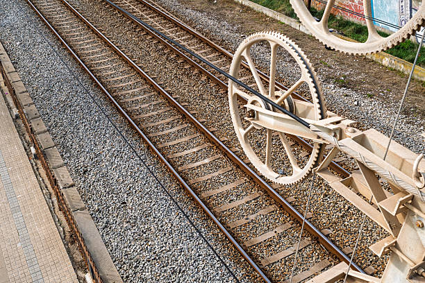 Overhead view of railway tracks with gravel ballast beside a station platform, featuring large industrial pulley wheels and cables in the foreground.