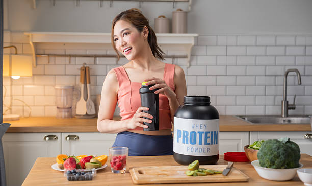 Asian fitness woman with whey whey protein container on kitchen table with fresh organic vegetables.