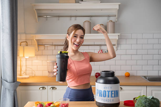 Smiling Asian fitness woman showing shaker bottle and flexing biceps with whey protein container and healthy vegetables in kitchen