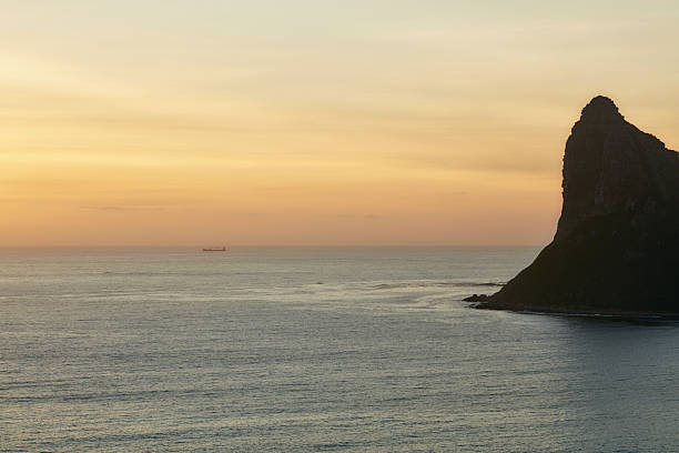 A serene coastal scene at sunset shows calm waters, a rugged rock formation to the right, and a distant ship on the horizon. The golden hour sky blends warm hues of orange and gold, evoking peaceful, travel-inspired moods and the beauty of South Africaâs Western Cape coastline. Hout Bay
