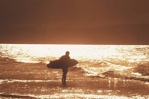 Silhouette of a surfer walking along the shoreline carrying a surfboard at golden hour sunset, with warm orange light reflecting on wet sand and gentle waves. Scenic coastal landscape with distant mountains in the background, evoking adventure and relaxation