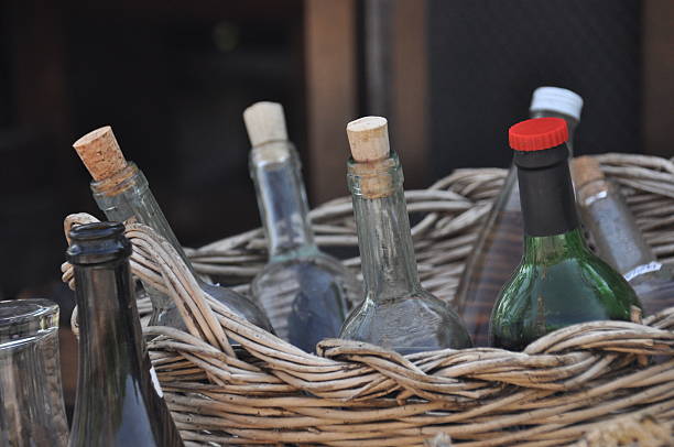 A woven basket filled with assorted glass bottles placed in a natural outdoor environment. Sunlight highlights the bottles, creating a warm atmosphere.