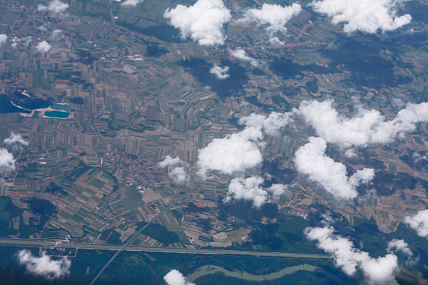 High altitude view of countryside with roads weaving through geometric fields. A river curves at the bottom right, partly veiled by scattered clouds