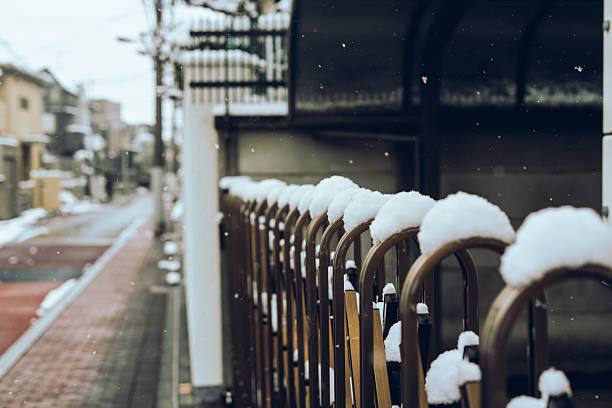 A close-up winter scene of a snow-capped accordion-style retractable gate along a quiet street in Japan. Falling snow and shallow depth of field create a calm seasonal mood, suitable for concepts such as winter weather, residential security, and everyday life.