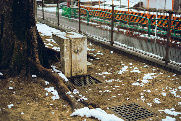 A public park drinking fountain stands in a snowy landscape with patchy melting snow on the ground. Suitable for themes such as winter season, cold weather, public facilities, outdoor parks, and seasonal transition.