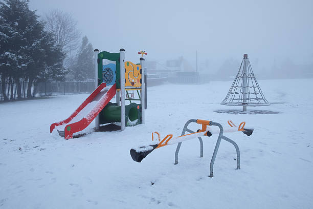 An empty playground covered in snow and obscured by fog or heavy mist. Colorful equipment (slide, seesaw, climbing net) contrasts with the cold, atmospheric winter day.