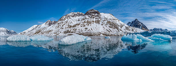 This stunning Arctic panorama captures the pristine beauty of Magdalenefjorden in Spitsbergen, Svalbard, Norway. The image showcases towering, snow-covered mountain peaks perfectly mirrored in the calm, deep blue waters of the fjord. Scattered ice floes and small ice fragments float on the surface, emphasizing the raw, untouched nature of the polar region. The vibrant blue sky and sharp contrasts highlight the dramatic textures of the rock and snow. Ideal for travel editorials, environmental awareness campaigns, and nature-themed commercial projects. The scene evokes a sense of tranquility and the immense scale of the Arctic wilderness, making it a perfect visual for high-end travel agencies, climate research publications, or interior decor pieces focused on natural serenity.