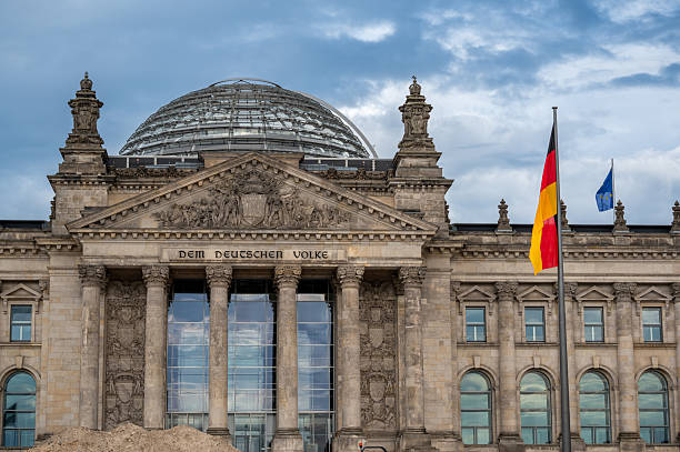 An angled architectural view of the Reichstag Building in Berlin, featuring ornate stonework, classical columns, sculptures, and the modern glass dome. The German and European flags wave above this historic seat of parliament on a cloudy spring day.