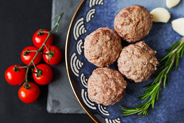 Four meatballs rest on a blue plate next to cherry tomatoes and fresh herbs, prepared for a meal.