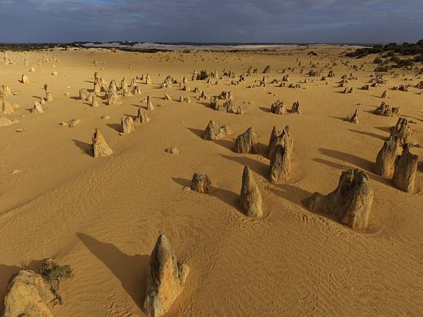 The pinnacles desert of Nambung National Park landscape with jagged stone pinnacles rising from warm sand beneath a expansive, partly cloudy sky, evoking solitude, rugged beauty, and timeless geology.