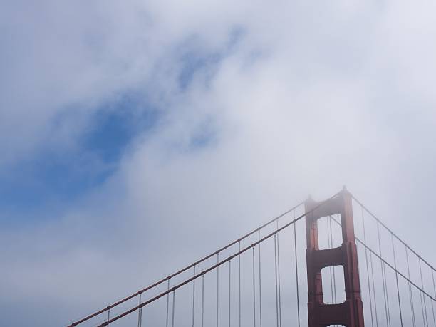 View of the Golden Gate Bridge partially obscured by fog, seen through a chain-link fence in San Francisco, California. The image captures the city’s iconic landmark in its characteristic misty atmosphere, symbolizing both beauty and mystery along the Pacific coast.