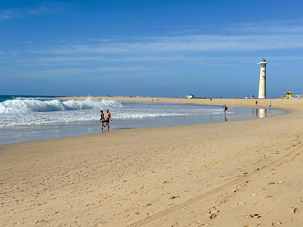 The striking red and white Far del Fangar lighthouse stands tall amid the rolling sand dunes of Delta del Ebro, Catalonia, Spain. Surrounded by a vast arid landscape, the lighthouse serves as a beacon against the backdrop of distant mountains and wind turbines. The golden sand and soft lighting create a serene and almost surreal coastal desert scene, highlighting the contrast between nature and maritime architecture