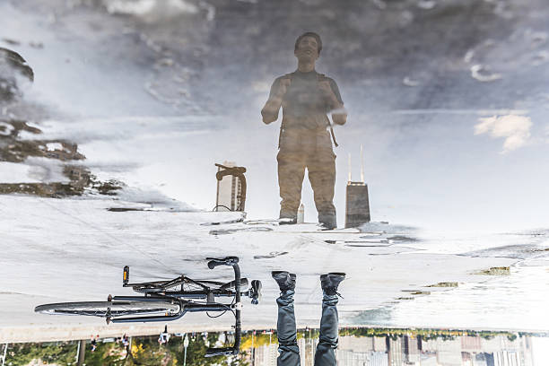 Man standing with bicycle, reflecting in a large puddle in an urban area, showing a clear upside down image of the Chicago skyline