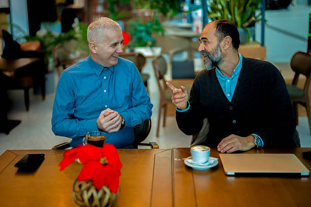 Two happy men laughing and having a casual conversation while sitting at a table in a modern cafe, enjoying coffee during a friendly business meeting or social gathering