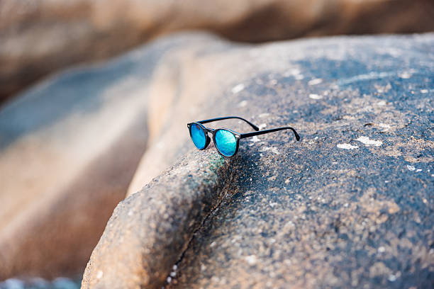 Stylish blue mirrored sunglasses are resting on a large weather-worn granite rock, evoking concepts of summer vacation, travel, and tropical beach leisure