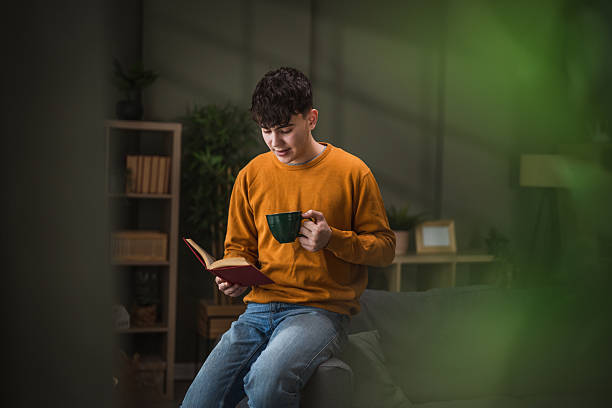 Young man enjoying a tranquil moment while reading a novel and holding a warm mug of coffee, sitting comfortably on a sofa in his cozy home living room
