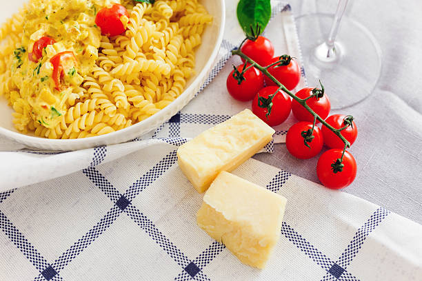 Pasta is placed in a bowl next to fresh tomatoes, cheese, and a glass on a cloth-covered table.