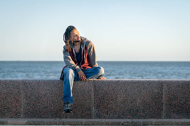 a young man with dreadlocks, dressed in a hoodie and jeans, is sitting on a wall, looking thoughtfully out to sea