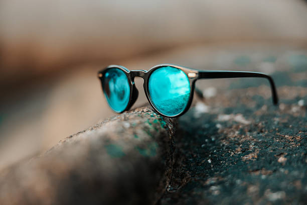 Stylish sunglasses with vibrant blue mirrored lenses reflecting the sparkling ocean and sky, resting on a textured rock, symbolizing a relaxed tropical lifestyle and travel vacation