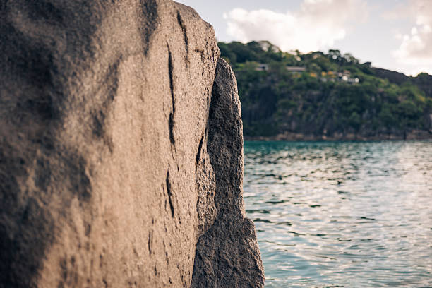 Jagged granite rock framing a turquoise ocean view with lush green hills under a bright sky, creating a serene and timeless natural landscape on an island