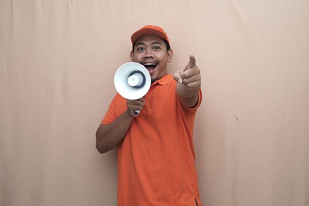 Southeast Asian courier in orange uniform shouting into a megaphone and pointing upward, promoting an announcement or marketing message on beige background.