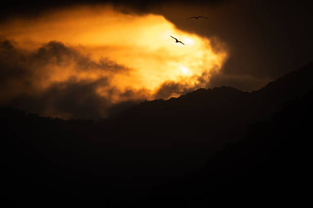 A dramatic landscape scene showing dark mountain silhouettes in the foreground with thick clouds partially opening to reveal a glowing orange sky. Two birds are visible in flight against the bright opening in the clouds, adding scale and motion.