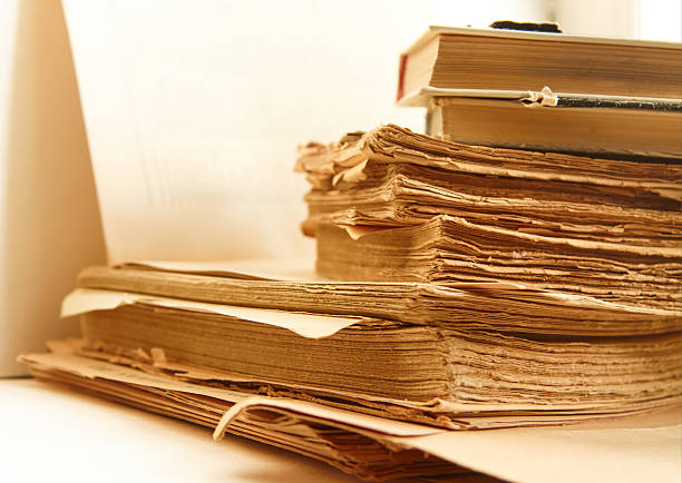 Aged books with yellowed pages and worn covers are stacked on a table, accompanied by loose sheets of paper and soft ambient lighting