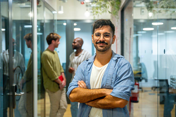 Young man smiling at the camera, standing with crossed arms in a modern coworking office space while colleagues are networking and collaborating in the blurred background