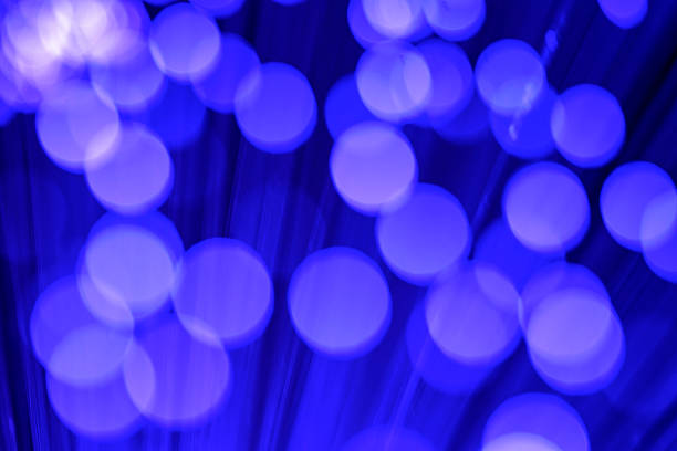Close-up view of optical fiber cables glowing with blue light against a solid black background, creating an abstract pattern of soft, circular bokeh highlights and bursts of brighter illumination. The image captures the essence of data transmission and digital connectivity through luminous strands that evoke streams of information flowing in a high-tech environment. Ideal for illustrating concepts in telecommunications, internet infrastructure, big data, AI, and futuristic innovation in business and science publications. The horizontal composition emphasizes the complexity and beauty of fiber optic technology without any people or identifiable objects, focusing on the ethereal, glowing orbs that suggest speed and global communication networks. Suitable for editorial use in articles on digital transformation, cybersecurity, and engineering advancements.