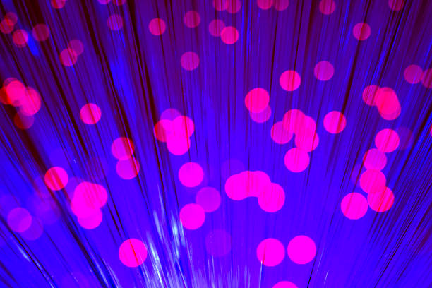 Close-up view of optical fiber cables glowing with blue light against a solid black background, creating an abstract pattern of soft, circular bokeh highlights and bursts of brighter illumination. The image captures the essence of data transmission and digital connectivity through luminous strands that evoke streams of information flowing in a high-tech environment. Ideal for illustrating concepts in telecommunications, internet infrastructure, big data, AI, and futuristic innovation in business and science publications. The horizontal composition emphasizes the complexity and beauty of fiber optic technology without any people or identifiable objects, focusing on the ethereal, glowing orbs that suggest speed and global communication networks. Suitable for editorial use in articles on digital transformation, cybersecurity, and engineering advancements.