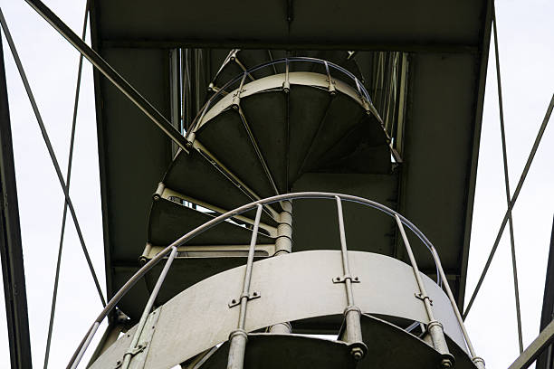 The upward viewpoint turns the stair into a graphic spiral, highlighting bolts, rails, and repeating forms. Cool metallic tones keep the focus on shape and engineering. This image suits themes of architecture, progress, and structural design.