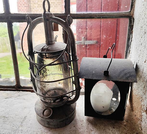 A weathered metal oil lamp with a glass globe sits next to a black rectangular candle lantern. Both objects are covered in a thick layer of dust and grime from long-term storage or neglect. The lanterns are positioned on a rough white ledge in front of an old window with a red door visible outside