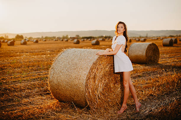 Young adult woman standing barefoot next to a hay bale in a golden field during sunset