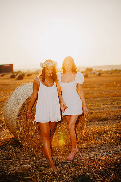 Two young adult women walking barefoot in a field holding hands at golden hour
