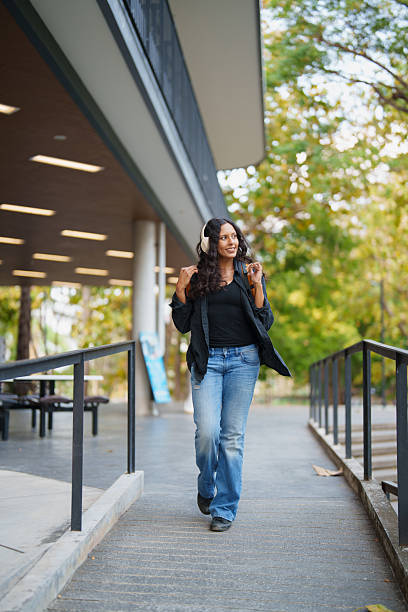 Young woman walking through a modern university campus with headphones, smiling and enjoying musiccasual student style, backpacked, relaxed and confident in warm natural light