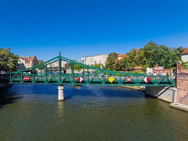 Pedestrian Bridge Over Odra River in Wroclaw, Poland. Pedestrian bridge crossing the Odra River in Wroclaw, Poland. Aerial view showing a green steel bridge, riverbanks, park area, and surrounding urban architecture on a sunny day. European city scene highlighting transportation, travel, and city infrastructure.