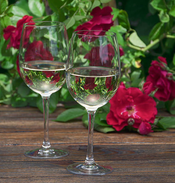 Two elegant glasses filled with a clear refreshing summer drink, placed on a wooden table outdoors, surrounded by flowers and greenery. The image represents hydration, relaxation, natural freshness and a light, healthy lifestyle during a warm sunny day.