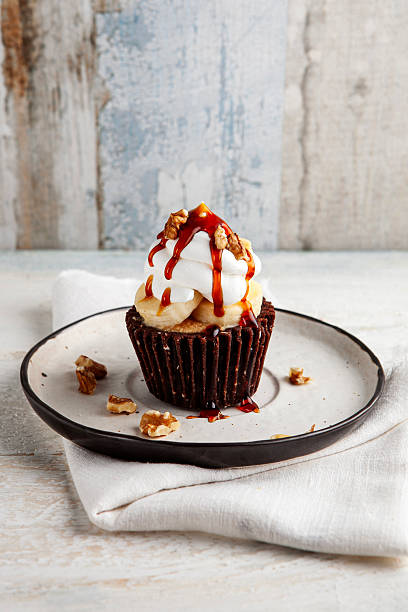 Delicious homemade chocolate muffin topped with fresh banana slices, fluffy whipped cream, walnuts, and sweet caramel sauce on a rustic plate.
