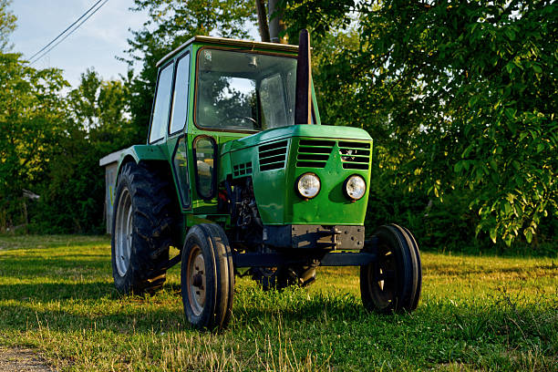 A green agricultural tractor sits on a grassy field surrounded by trees, illuminated by warm afternoon light. Its large rear tires, enclosed cab, and rugged design highlight classic farming equipment