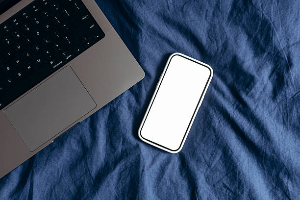 Flat lay of a smartphone with blank white screen and laptop on a blue bed, representing remote work, digital lifestyle, home office, and modern technology mockup with copy space.