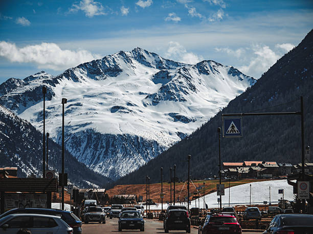 A stunning view of a massive, snow-covered mountain peak looming over a busy street in a European alpine village. The contrast between daily urban life with cars and the grand scale of nature creates a breathtaking scene.