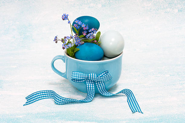 Blue color Easter eggs in light blue color cup with ribbon decorated with forget-me-not  natural blossoms, vibrant colored Easter composition on light blue background, Easter holiday table decoration, copy space
