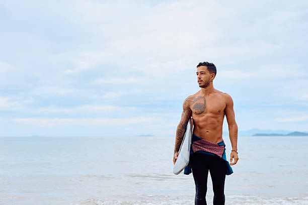 Muscular man with tattoos standing on beach, holding a surfboard, looking intensely at the calm cloudy ocean