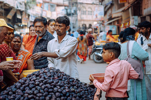 Varanasi, India - November 3, 2024:  Vibrant street scene in Varanasi, India, showing a crowded market stall piled with plums, diverse shoppers, vendors, and a child with a basket, capturing daily life and commerce.