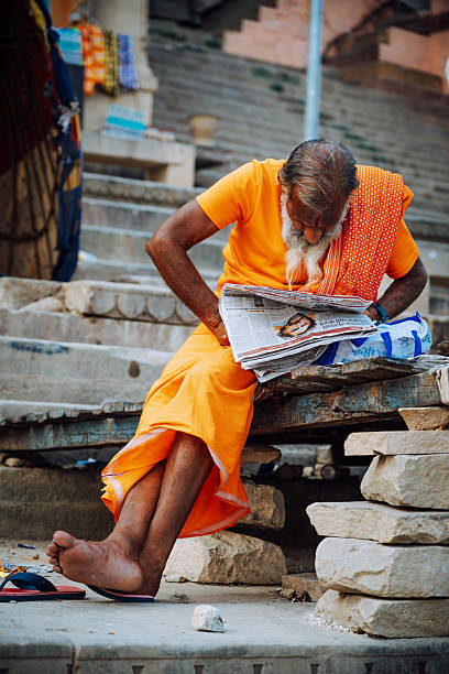 Varanasi, India - November 3, 2024:  A weathered sadhu in a bright orange robe sits on stacked stones in Varanasi, India, reading a newspaper. The scene conveys calm devotion, street life, and everyday ritual along the riverbank.