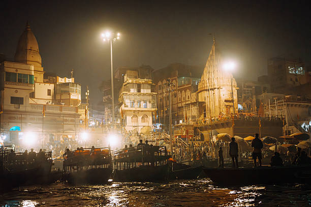 Vibrant night scene along the Ganges in Varanasi with illuminated ghats, temple spires, and boats on the river. Warm lights reflect on water as crowds gather, creating a lively Indian panorama.