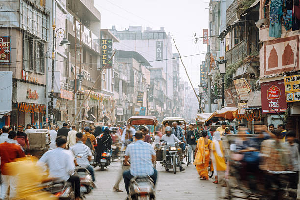 A crowded market street in Varanasi filled with scooters, pedestrians, rickshaws, and shopfronts. Colorful clothing and signage create a vibrant, chaotic urban atmosphere along busy old city lanes.