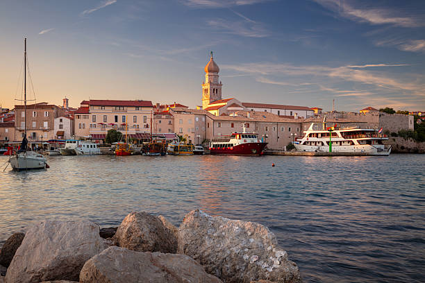 Krk, Croatia, cityscape at summer sunset. The skyline is dominated by Krk Cathedral, featuring its distinctive dome. The foreground shows a calm harbor with several boats docked, reflecting the gentle hues of the evening sky. Historic buildings line the waterfront, their warm tones highlighted by the setting sun. Rocky shoreline in the immediate foreground adds texture. The serene atmosphere captures the charm of this coastal town on Krk Island.